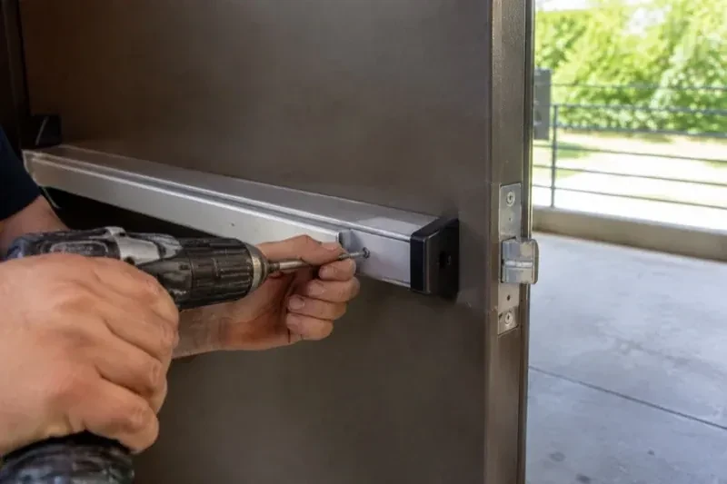 Hands using a power driver at the end of a horizontal push bar on an open metal door. — Panic & Exit Hardware, Mississauga